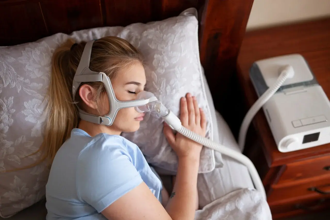 Mulher dormindo de lado com máscara de CPAP conectada ao aparelho sobre a mesa de cabeceira